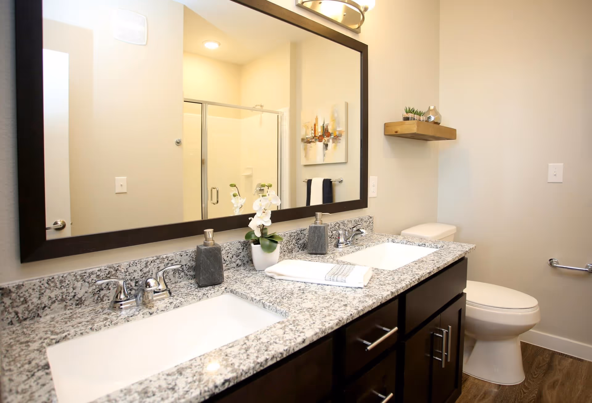 Bright modern bathroom with a double-sink granite vanity, large framed mirror, toilet, and a shower reflected in the mirror.
