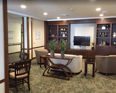 A cozy living room area with a beige sofa and two armchairs arranged around a wooden coffee table with two potted plants. Behind the seating area is a large wooden built-in shelving unit with a flat-screen TV mounted in the center and decorative items on the shelves. To the left, there is a small dining table with chairs. The room has recessed ceiling lights and a carpet with a leafy pattern.