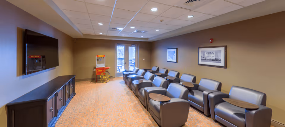 A cozy media room with two rows of comfortable gray armchairs, each equipped with a small wooden table. A large flat-screen TV is mounted on the wall opposite the chairs. The room has warm beige walls, framed vintage-style pictures, and a popcorn machine near the glass doors that lead to an outdoor area.