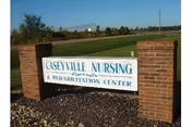 Outdoor view of a sign for Caseyville Nursing & Rehabilitation Center mounted between two brick pillars with grass and trees in the background under a clear sky.