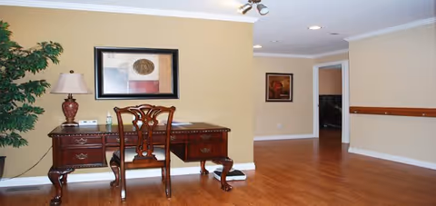 A spacious room with wooden flooring and beige walls featuring a classic wooden desk with a matching chair, a table lamp, and a framed abstract painting on the wall. There is a large green plant on the left side and an open doorway leading to another room in the background.