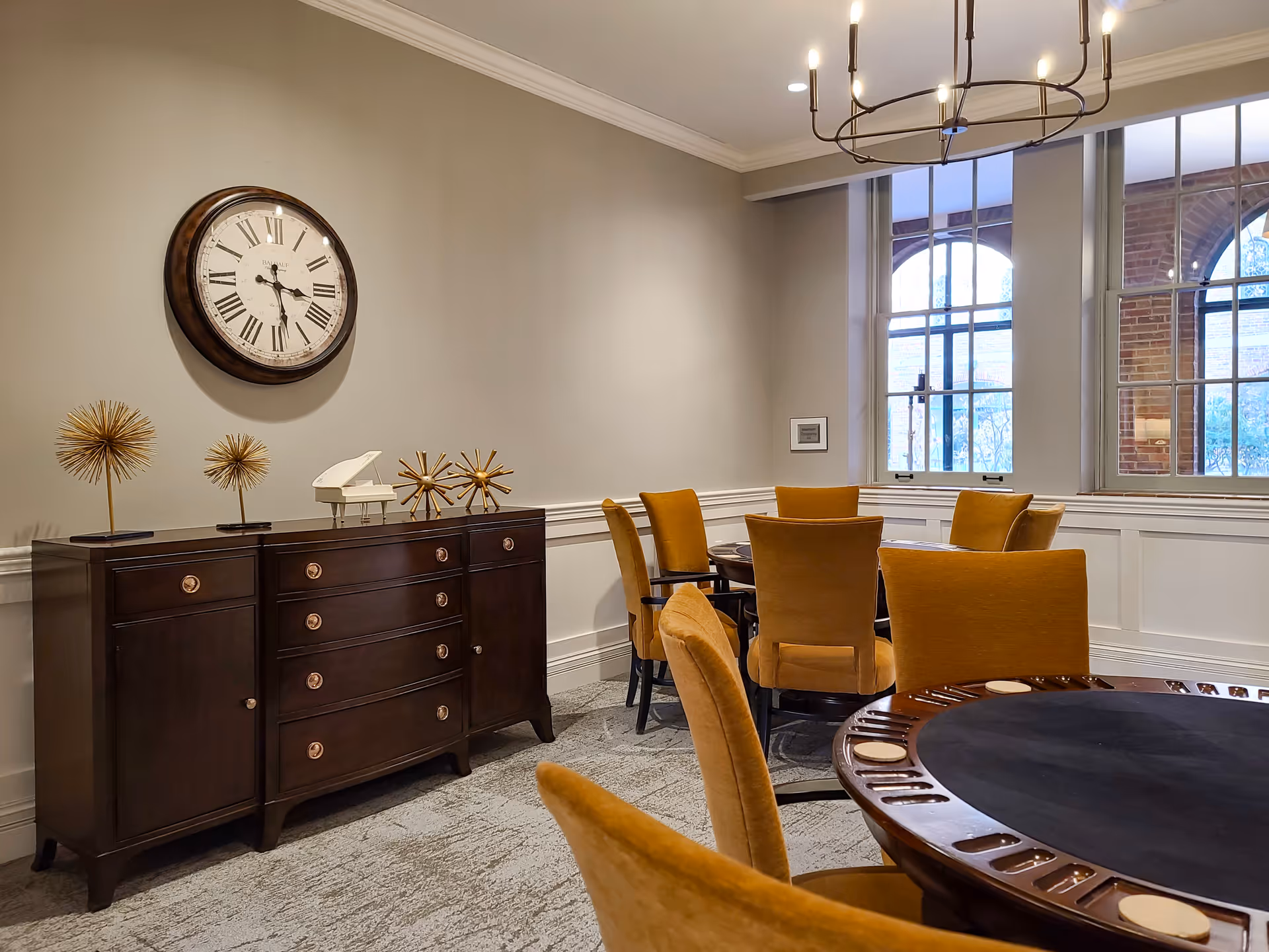A cozy interior room with a round table surrounded by mustard yellow upholstered chairs. A dark wooden sideboard with decorative items and a large wall clock with Roman numerals hangs above it. Large windows with arched brick frames let in natural light.
