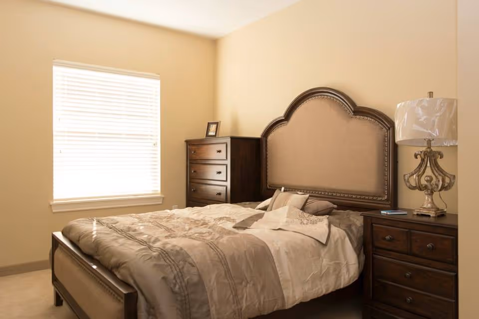 Well-lit bedroom with a large upholstered headboard, wooden nightstands and dresser beside a window with blinds.