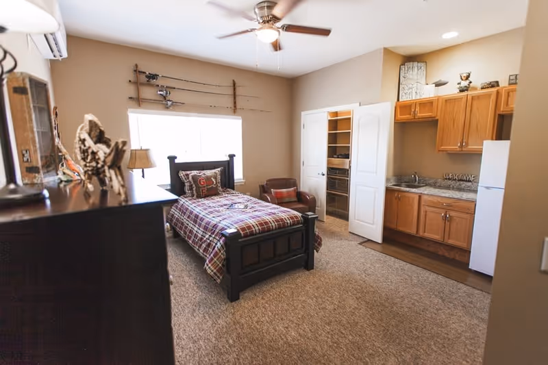 A cozy senior living room featuring a single bed with plaid bedding, a brown armchair with a red plaid pillow, a wooden dresser with decorative items, a ceiling fan with light, and a small kitchenette with wooden cabinets, a sink, and a white refrigerator. There is a window with fishing rods mounted above it and an open closet with shelves.