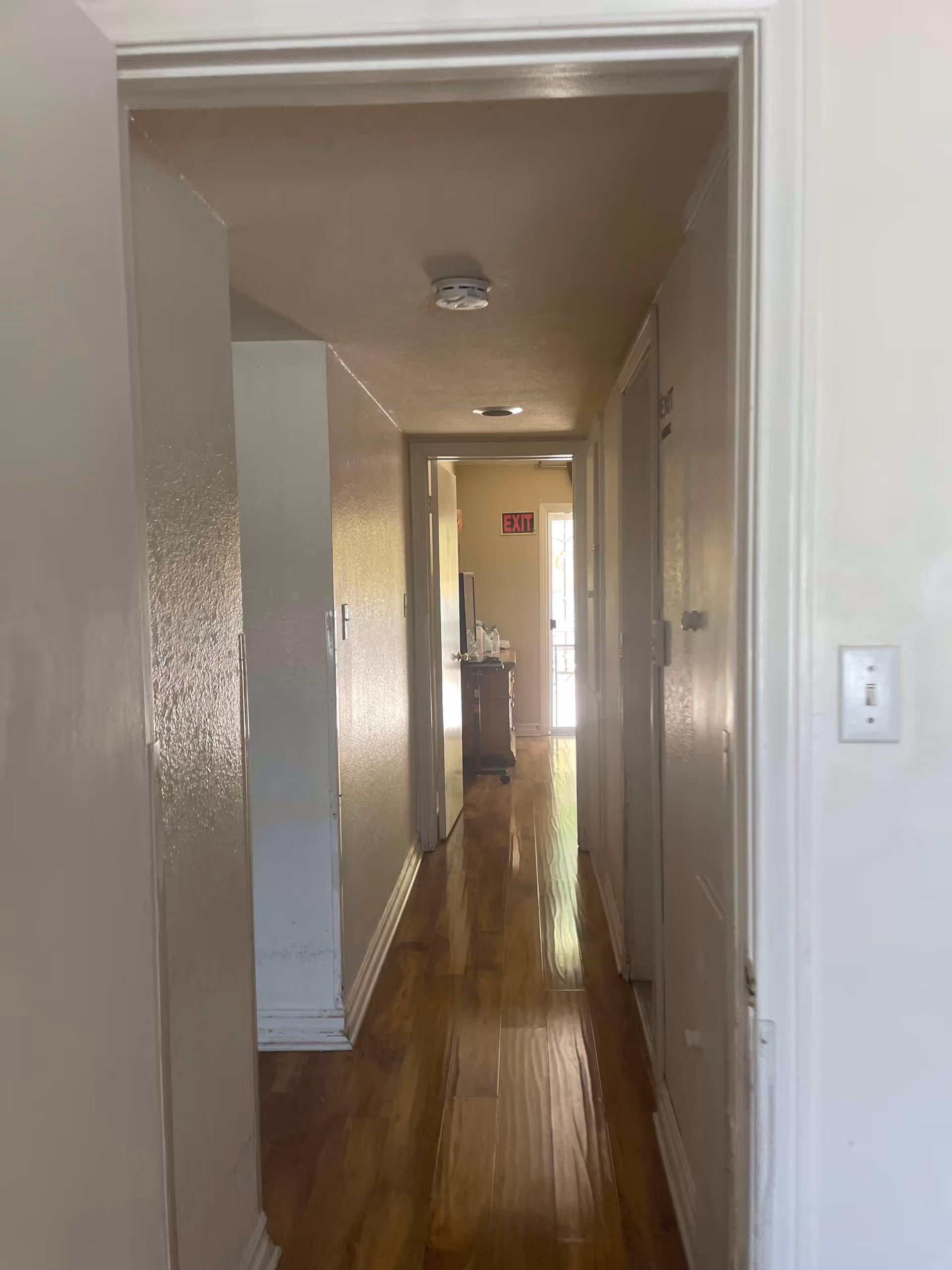 A narrow hallway with wooden flooring and cream-colored walls. There are doors on the right side and an open doorway on the left. At the end of the hallway, there is a small table with items on it and a visible exit sign above a door leading outside.