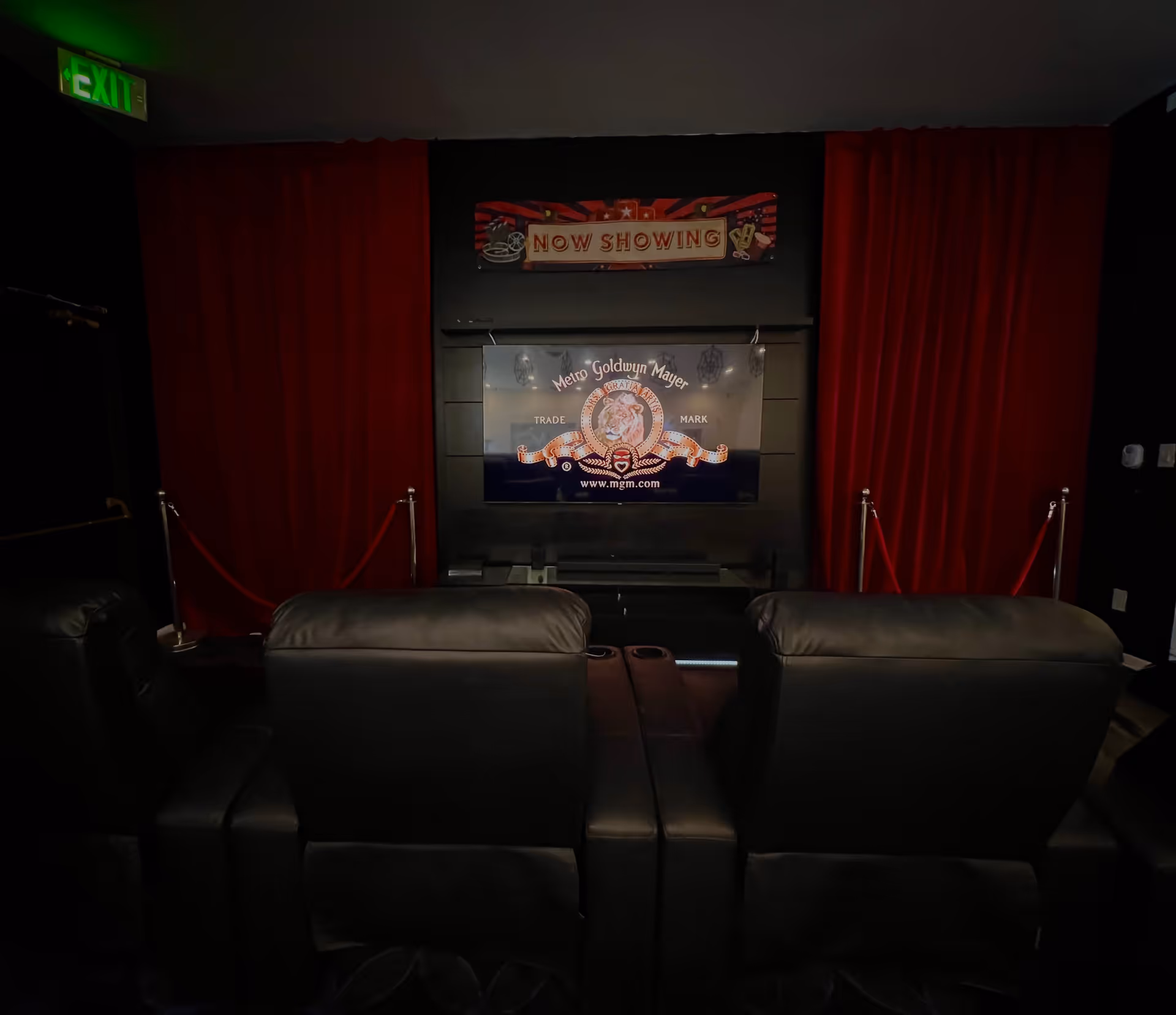 A small movie theater room with black leather recliner chairs facing a large screen displaying the Metro Goldwyn Mayer logo. The room has dark walls, red curtains on either side of the screen, and a 'Now Showing' sign above the screen. There is a green exit sign illuminated in the top left corner.