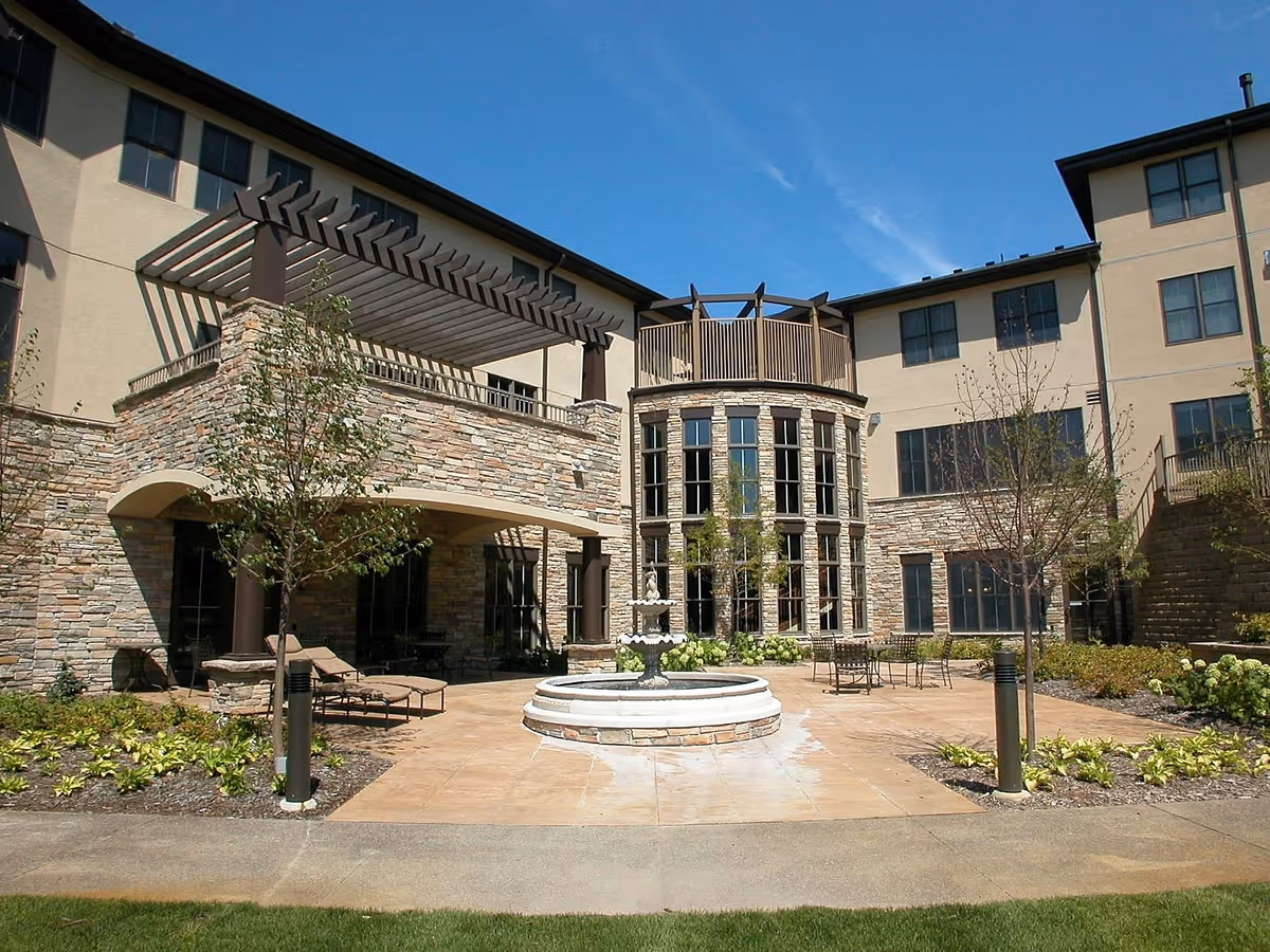 Outdoor courtyard area of a senior living facility with a stone fountain in the center, surrounded by patio seating, small trees, and landscaped plants. The building has a stone and beige exterior with large windows and a pergola on the upper level.