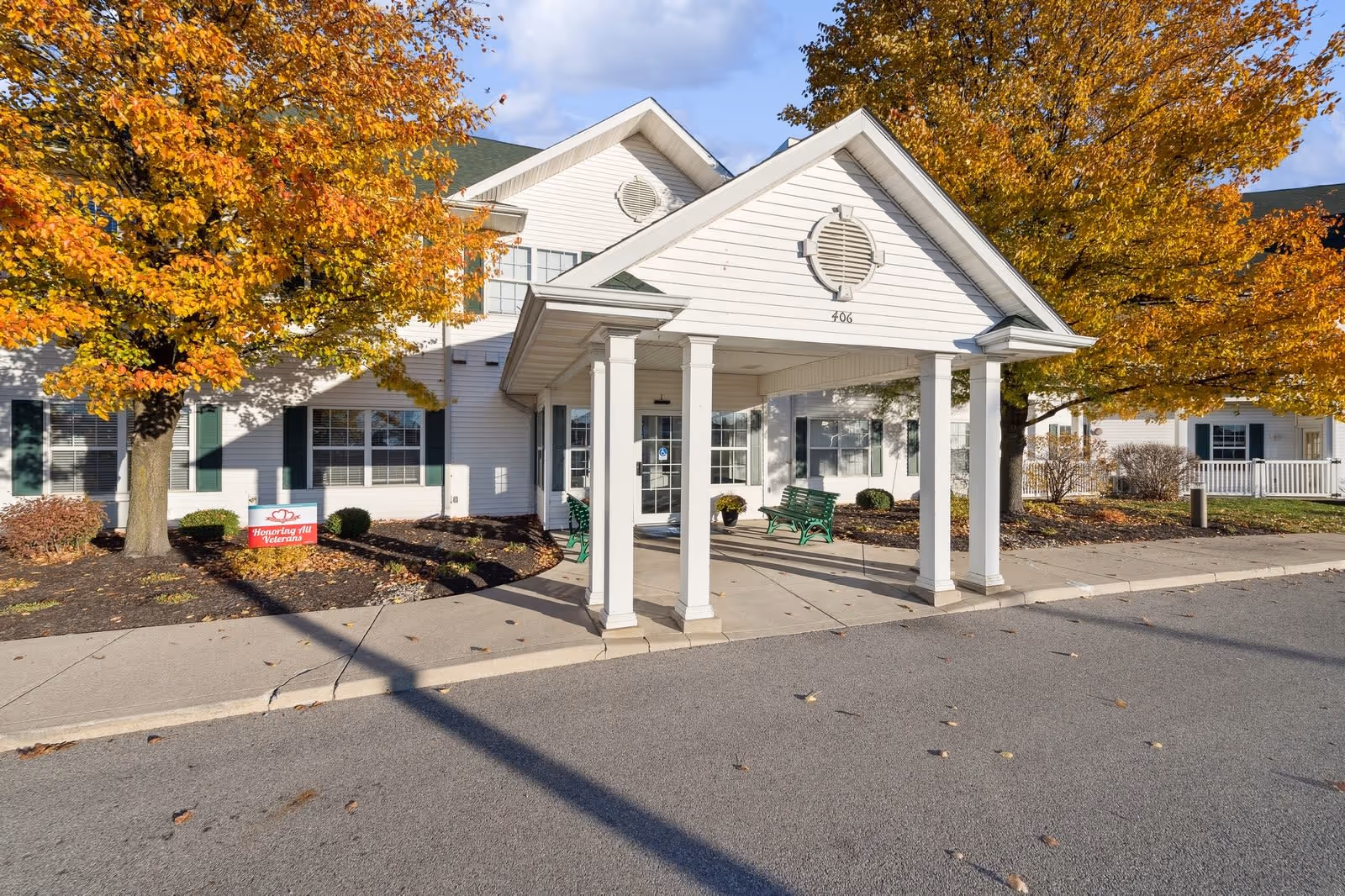 Exterior front entrance of a white senior living facility building with a covered porch supported by four white columns. There are green benches under the porch and autumn trees with orange and yellow leaves surrounding the building. A small sign near the tree reads 'Honoring All Veterans.'