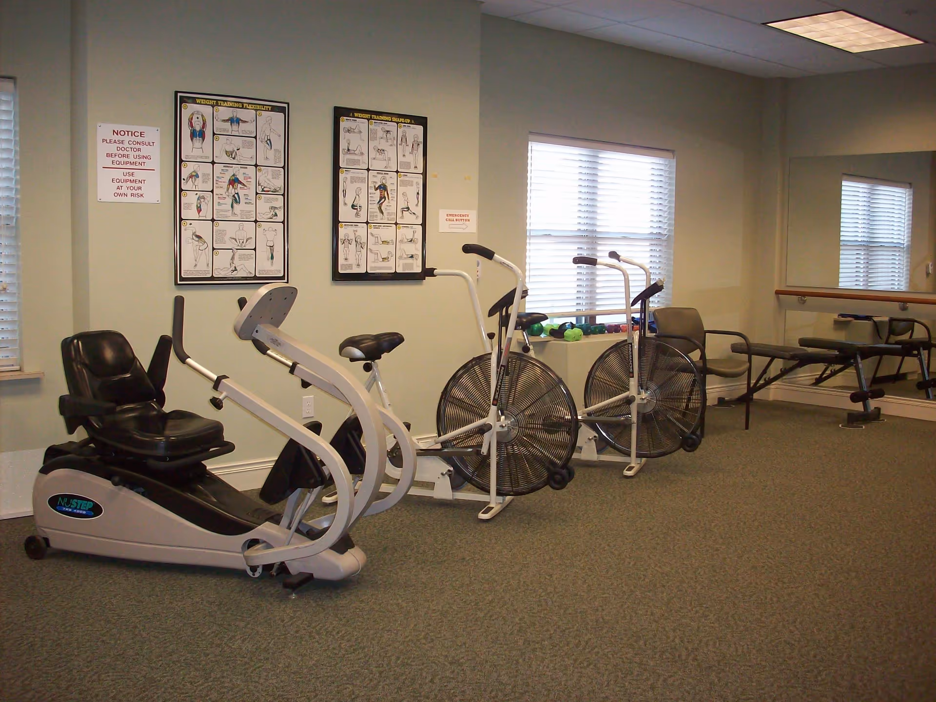 Interior view of a senior living facility exercise room with stationary exercise bikes, a recumbent bike, dumbbells on a windowsill, exercise posters on the wall, and a large mirror reflecting the room.