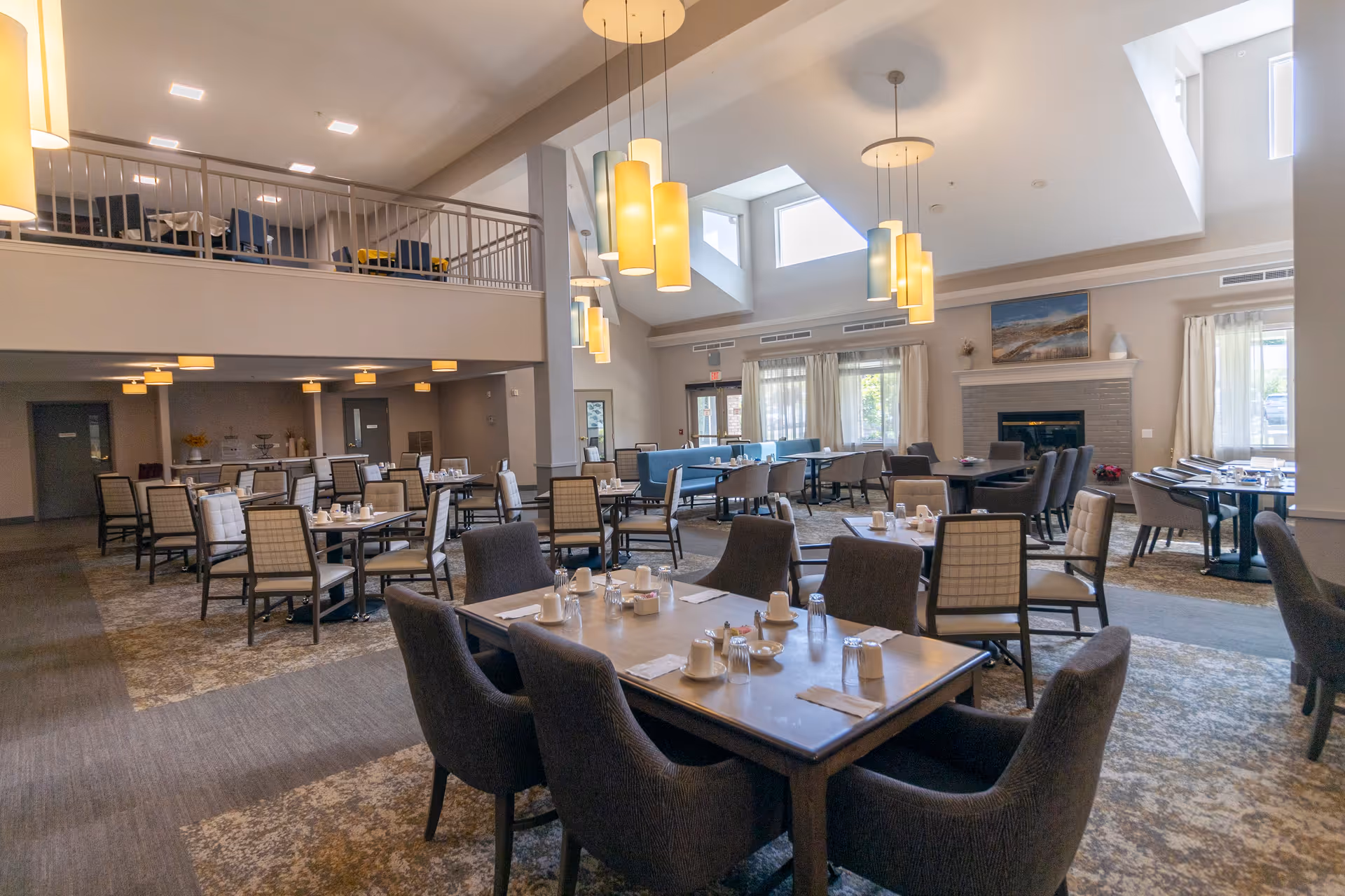 A spacious dining room with multiple tables and chairs arranged neatly. The room features high ceilings with modern hanging light fixtures, large windows allowing natural light, and a fireplace with a painting above it. The tables are set with cups, glasses, and napkins, ready for guests.