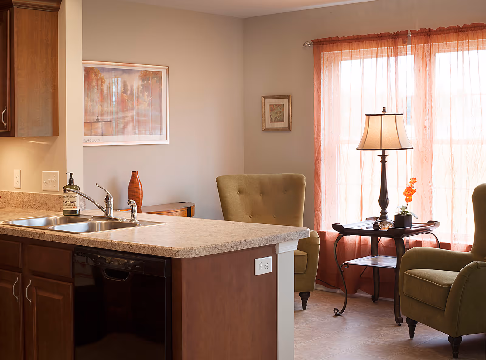 Open-plan interior showing a kitchen island with a sink in the foreground and a cozy seating area with two armchairs, side table and lamp by a window with sheer curtains.