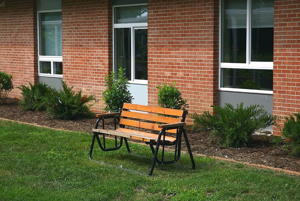 A wooden bench with black metal armrests and legs sits on a grassy lawn in front of a red brick building with several windows and small green shrubs along the building's foundation.