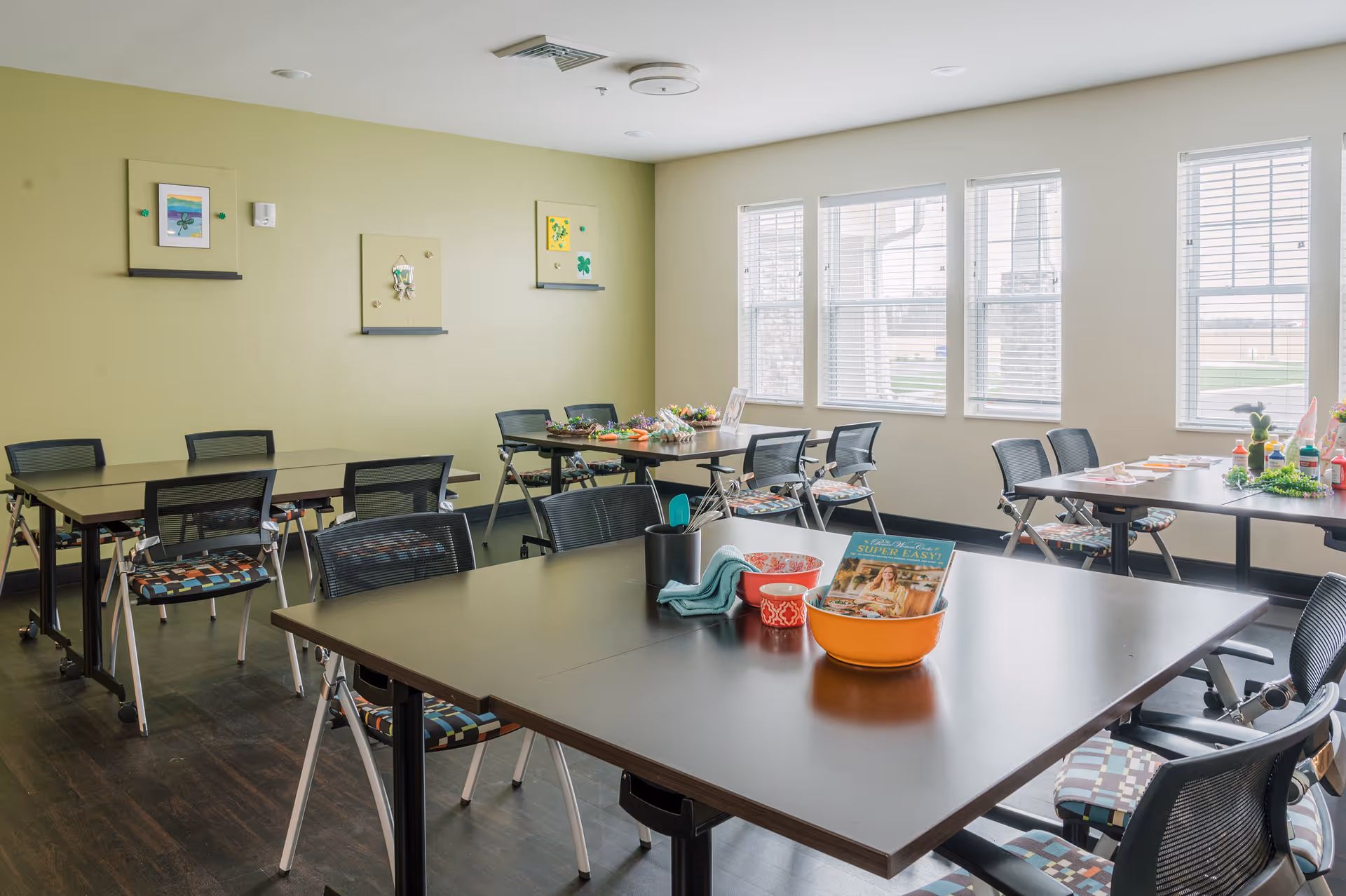 Bright room with multiple tables and chairs arranged for group activities or dining. The room has large windows with blinds letting in natural light, light green and white walls with framed artwork, and various items such as a cookbook, bowls, and utensils on the tables.