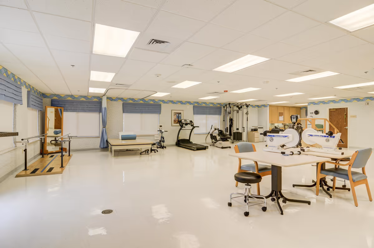A spacious physical therapy or rehabilitation room with exercise equipment including treadmills, stationary bikes, parallel bars for walking exercises, and tables with chairs. The room has a clean, bright interior with white floors, light-colored walls, and blue window valances.