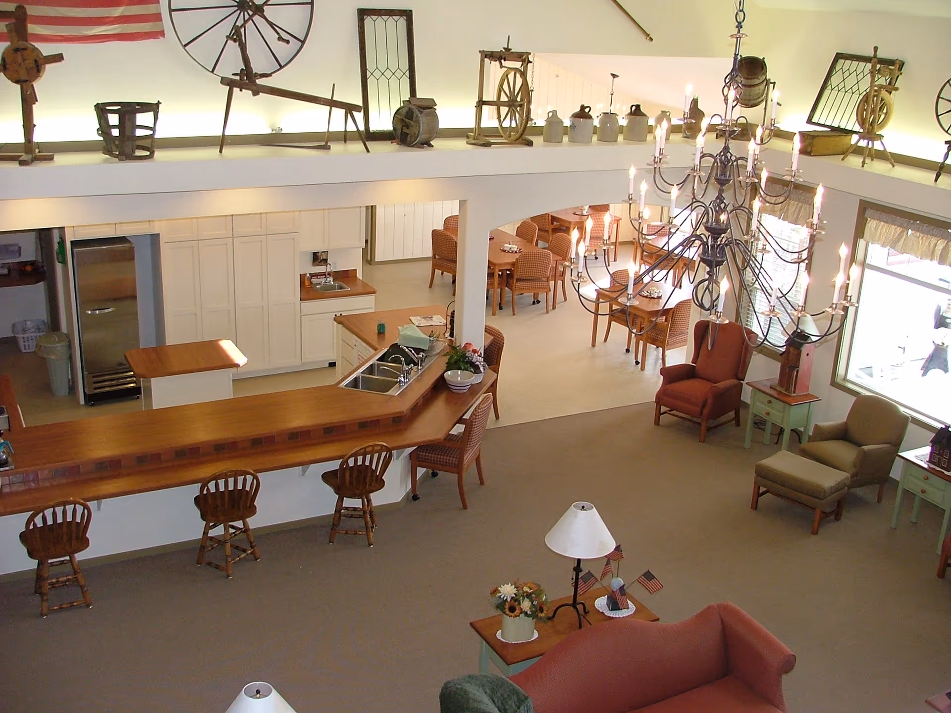 Open communal living and dining area with a long wooden service counter, seating, a large chandelier, and dining tables viewed from above.