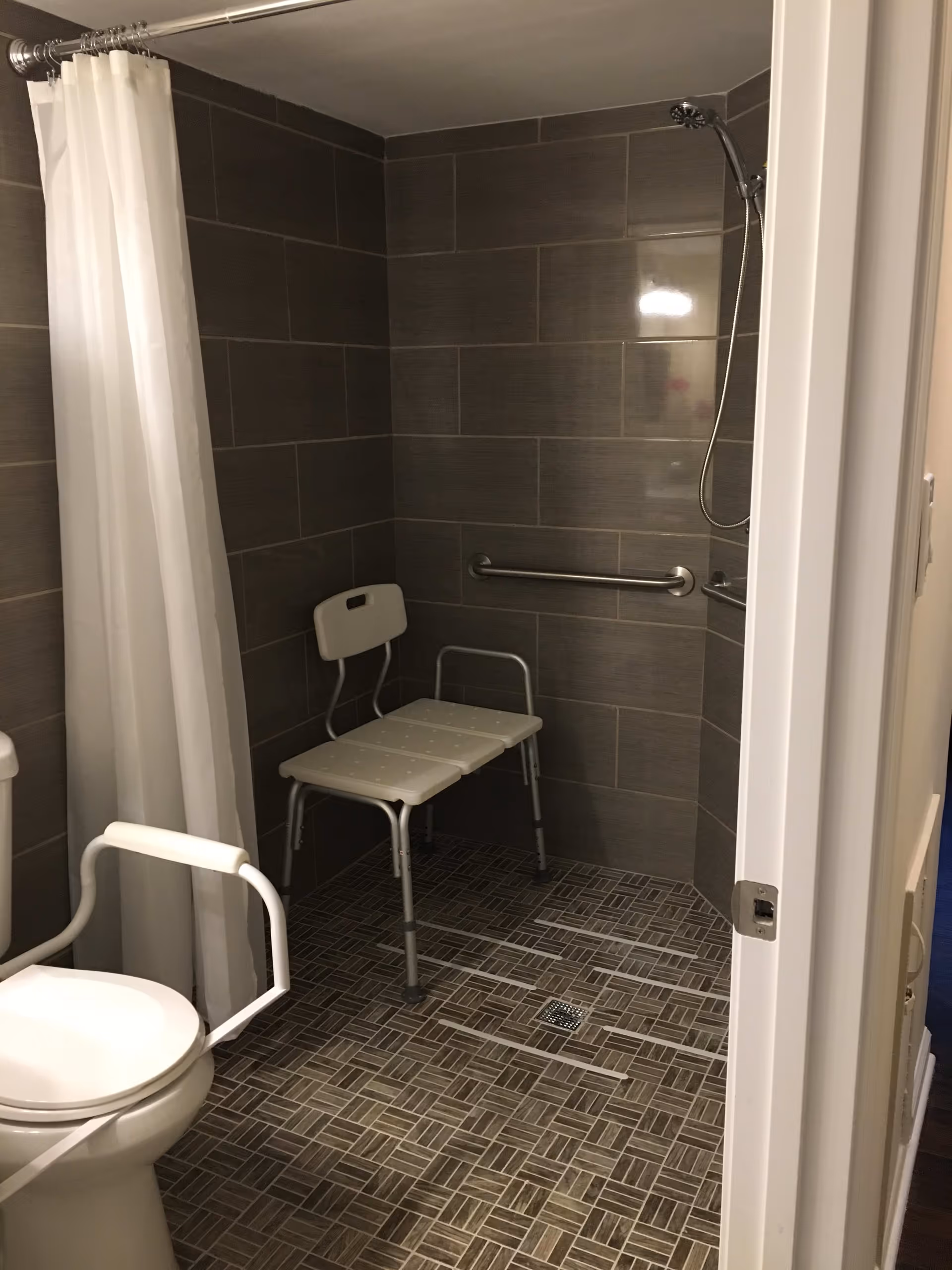 Accessible bathroom with a white toilet equipped with support rails, a walk-in shower area with a white shower chair, gray tiled walls, and a white shower curtain.