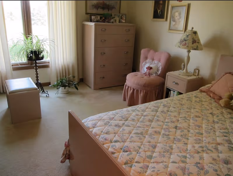 Light-filled bedroom with a floral quilted bed, pink upholstered chair, dresser and nightstand beside a window with plants.