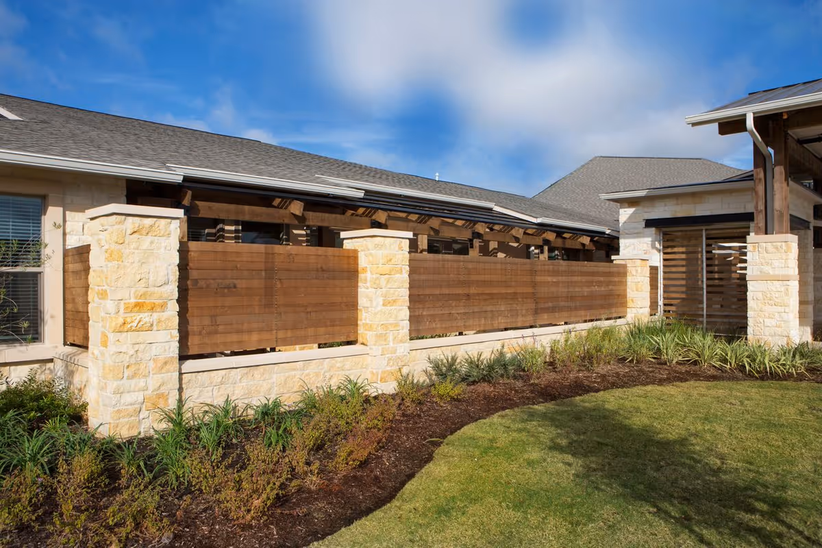 Exterior view of a senior living facility showing a stone and wood fence with landscaping in front and a building with a gray roof under a blue sky with some clouds.