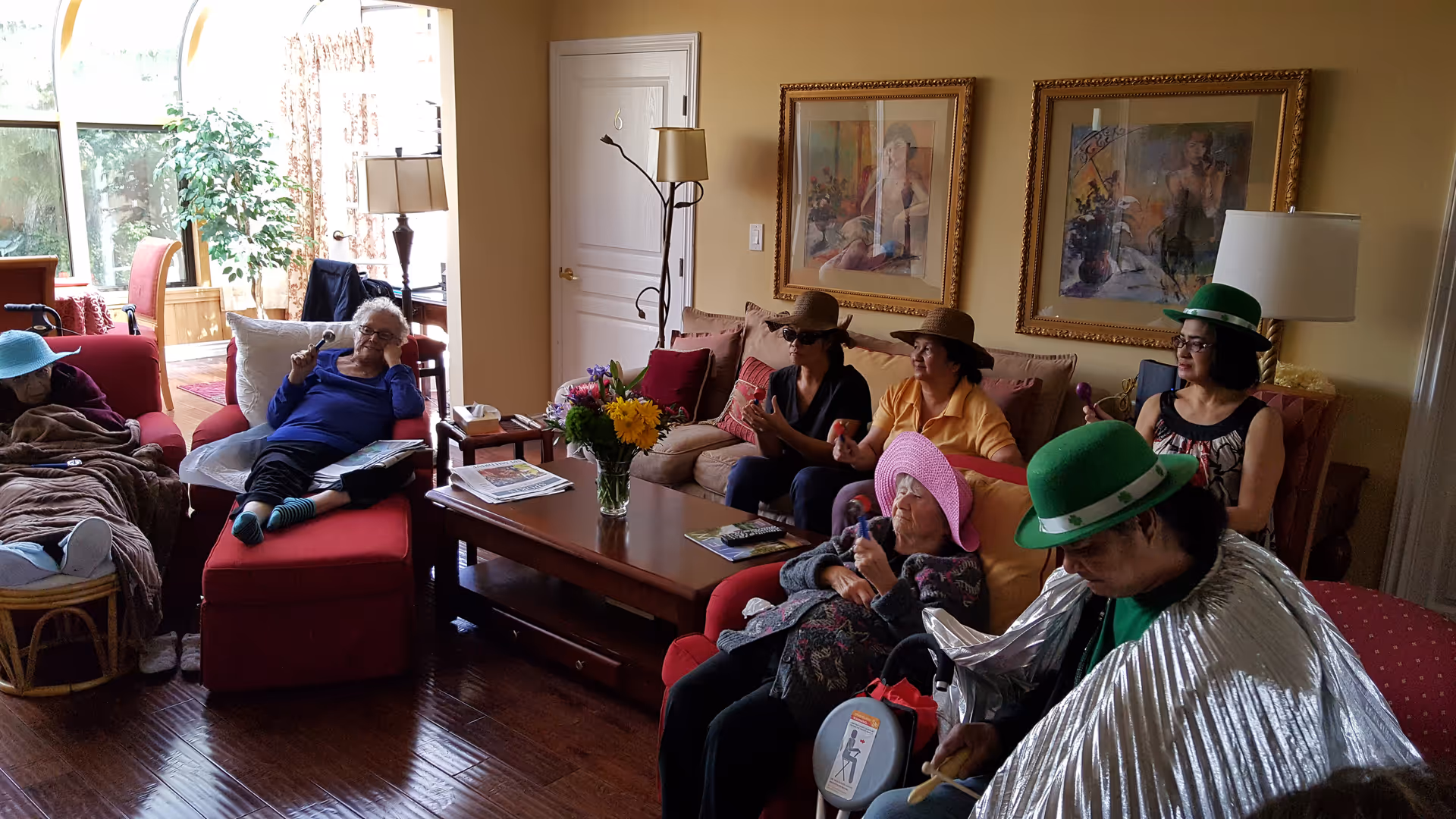 A group of elderly people sitting and relaxing in a cozy living room with wooden floors, colorful hats, and casual clothing. Some are seated on sofas and chairs, with a coffee table in the center holding a vase of flowers and magazines. The room has warm yellow walls, framed artwork, and a floor lamp.