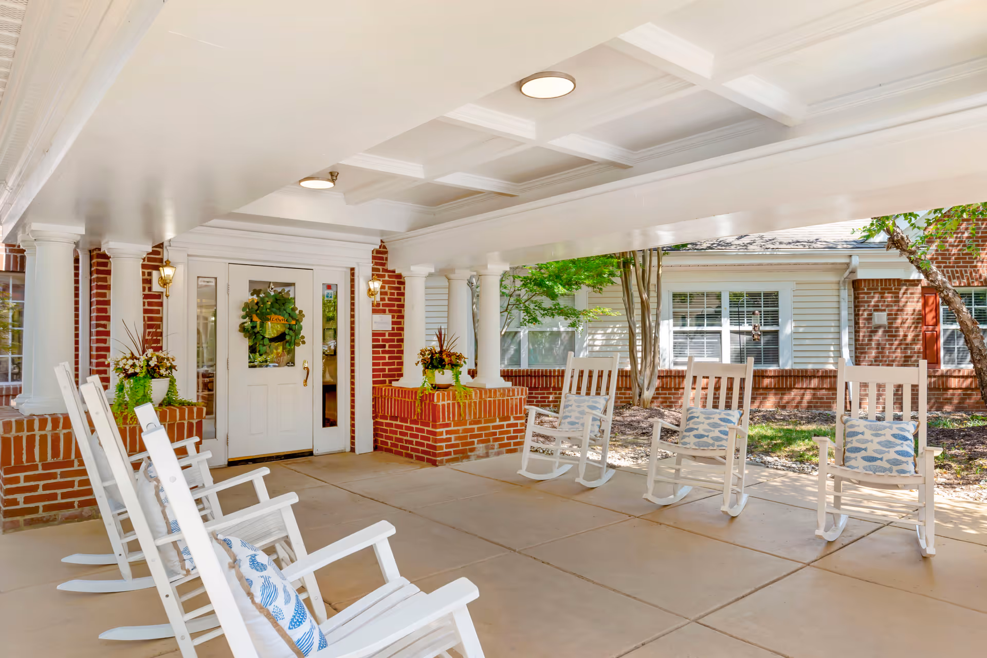 Covered outdoor patio area at Brookdale Cary with white rocking chairs arranged in two rows facing each other. The space features brick pillars and walls, white ceiling with recessed lighting, and potted plants near the entrance door decorated with a wreath. Trees and windows of the building are visible in the background.
