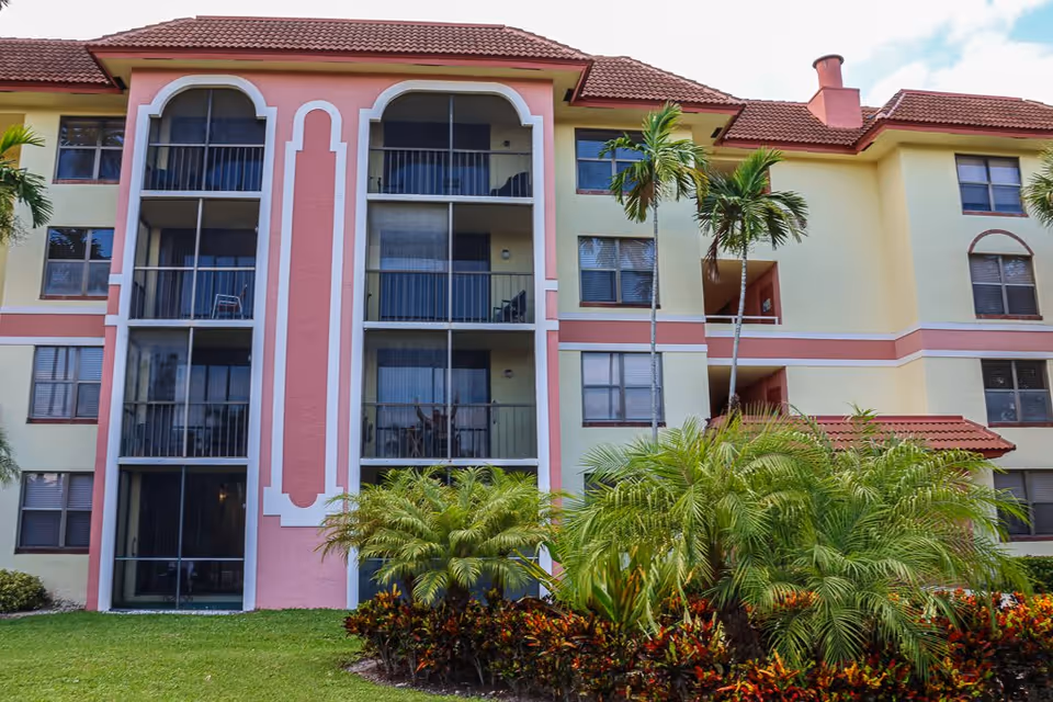 Front facade of a pastel-colored multi-story residential building with screened balconies, palm trees, and landscaped shrubs.