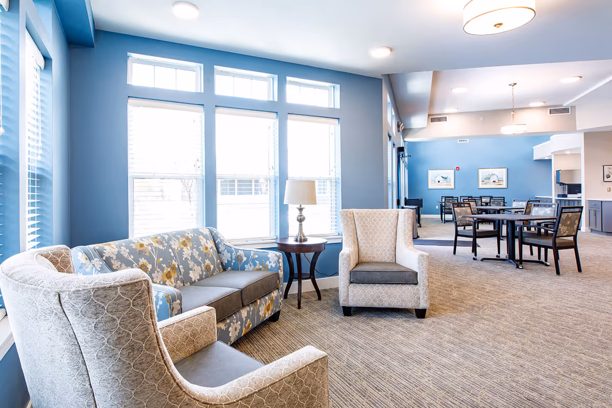 A bright and spacious common area with large windows letting in natural light. The room features a blue floral patterned sofa, two beige armchairs with patterned upholstery, and a small round wooden table with a lamp. In the background, there are several tables and chairs arranged for dining or activities, with blue walls and framed artwork.