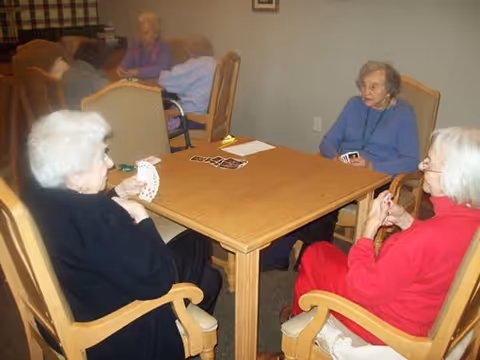 A group of elderly women sitting around a wooden table playing cards in a communal indoor setting with beige walls and cushioned wooden chairs.
