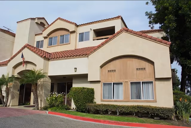 Exterior view of a two-story residential building with beige stucco walls and red tile roofing. The building has multiple windows, some with curtains, and a small balcony on the upper floor. There are palm trees, shrubs, and a well-maintained lawn in front of the building, along with a red-painted curb.