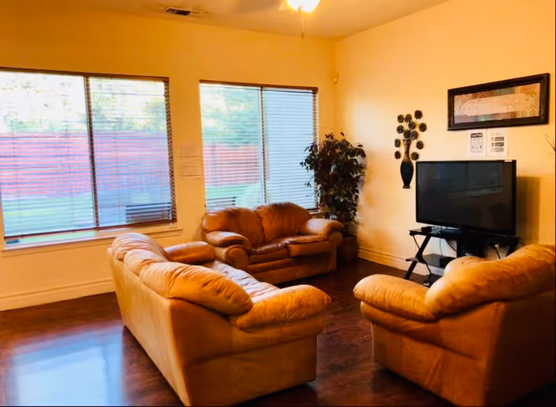 A cozy living room with three tan leather sofas arranged around a flat-screen TV on a black stand. The room has large windows with blinds, a potted plant in the corner, and a framed picture and decorative wall art above the TV. The floor is dark wood and the walls are painted a light color.