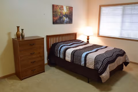 A simple bedroom with a single bed covered in a striped comforter in shades of brown, blue, and beige. Next to the bed is a wooden nightstand with a lamp on it. On the left side of the room is a wooden chest of drawers with three decorative vases on top. A window with blinds is on the right wall, and a framed picture of a forest scene hangs above the bed.