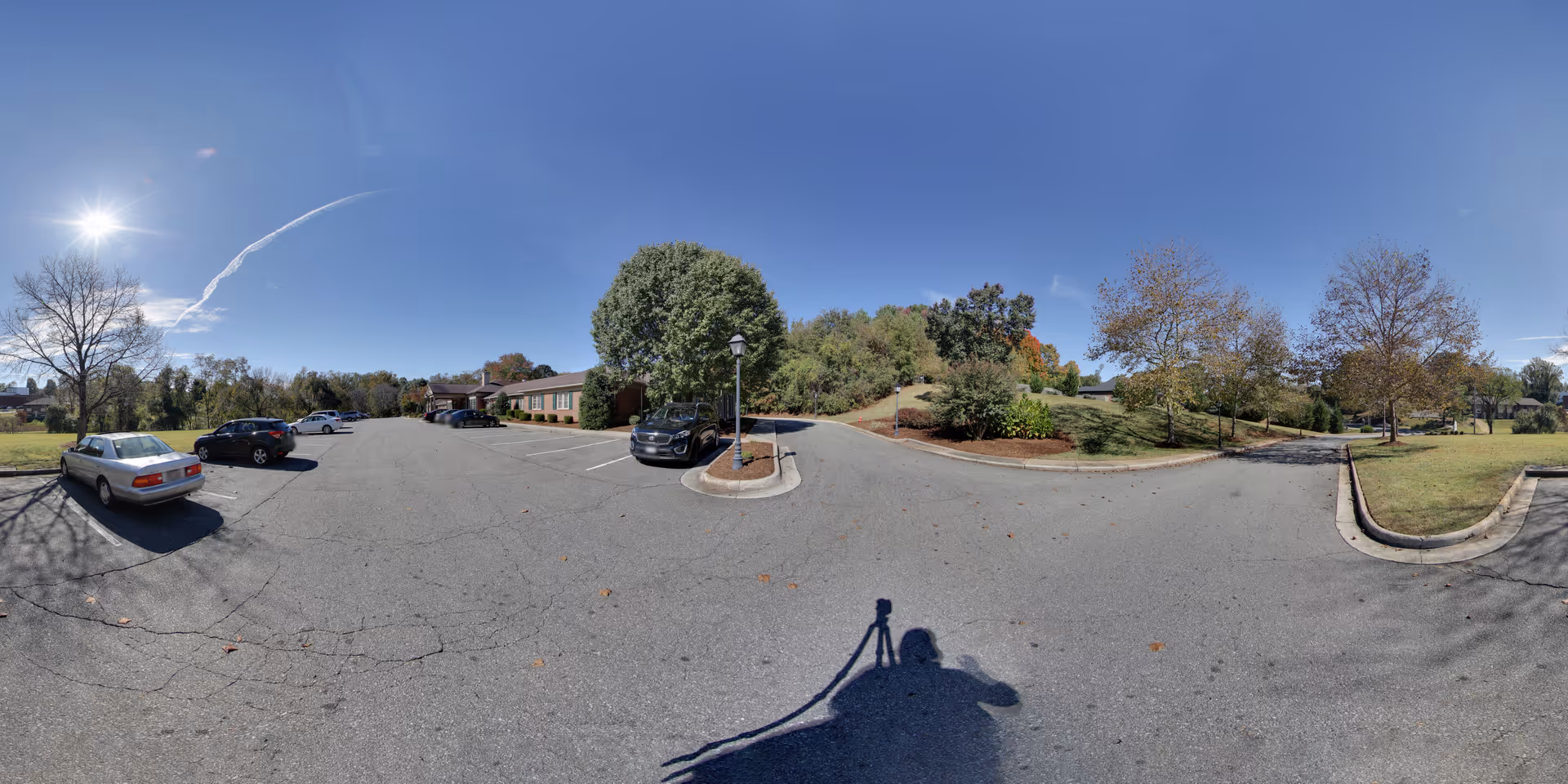 Wide panoramic view of a parking lot outside a single-story brick building with several parked cars. Trees with autumn foliage surround the area under a clear blue sky with the sun shining brightly.