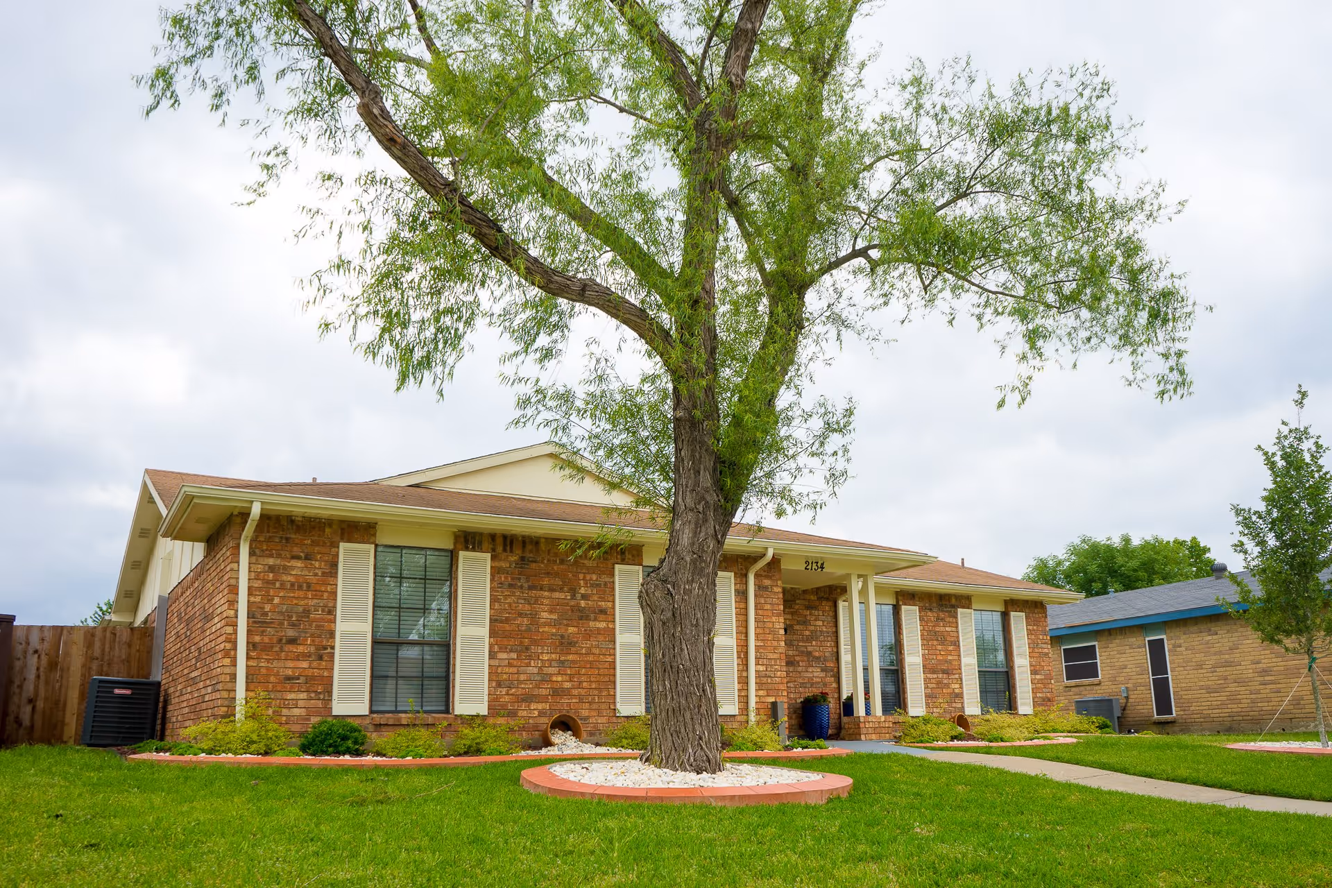 Single-story brick building with white shutters and a large tree in the front yard, surrounded by green grass and a concrete walkway leading to the entrance.