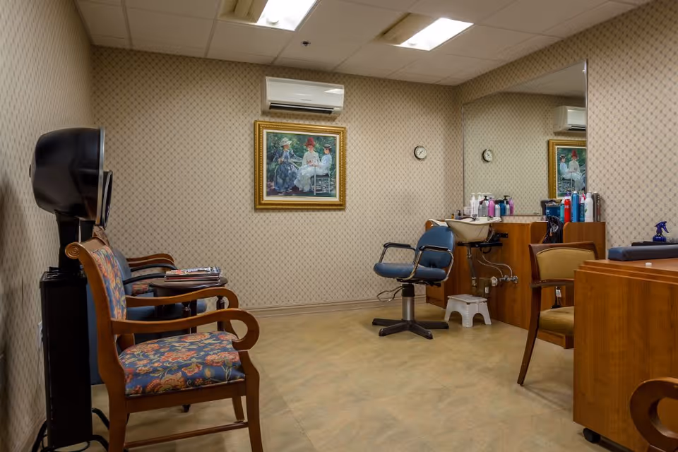 Interior of a small salon room with a hair washing station, a blue salon chair, wooden chairs with floral upholstery, a large mirror on the wall, a painting, and various hair care products on a counter.