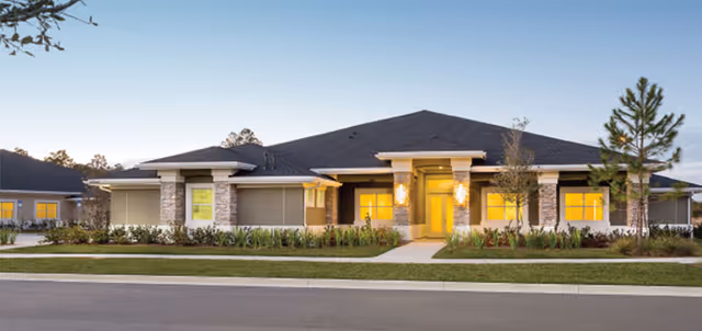 Single-story modern assisted living building front with a lit central entrance, walkway and landscaped lawn at dusk.