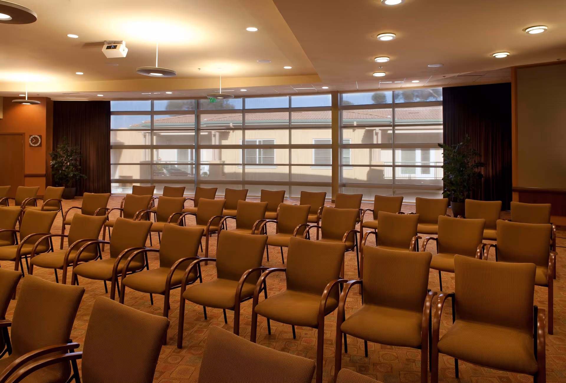 Empty meeting room with rows of upholstered chairs facing large windows and soft overhead lighting.