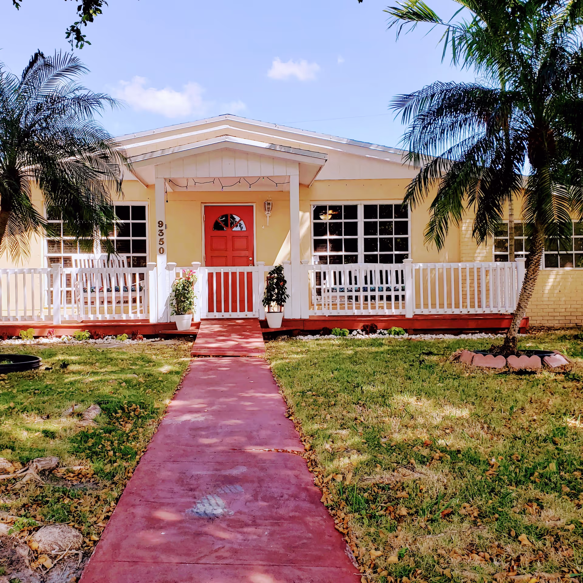 Front exterior view of a single-story yellow building with a red door, white railing, and a red walkway leading to the entrance. There are two potted plants on either side of the door, palm trees on both sides of the yard, and a clear blue sky with a few clouds.