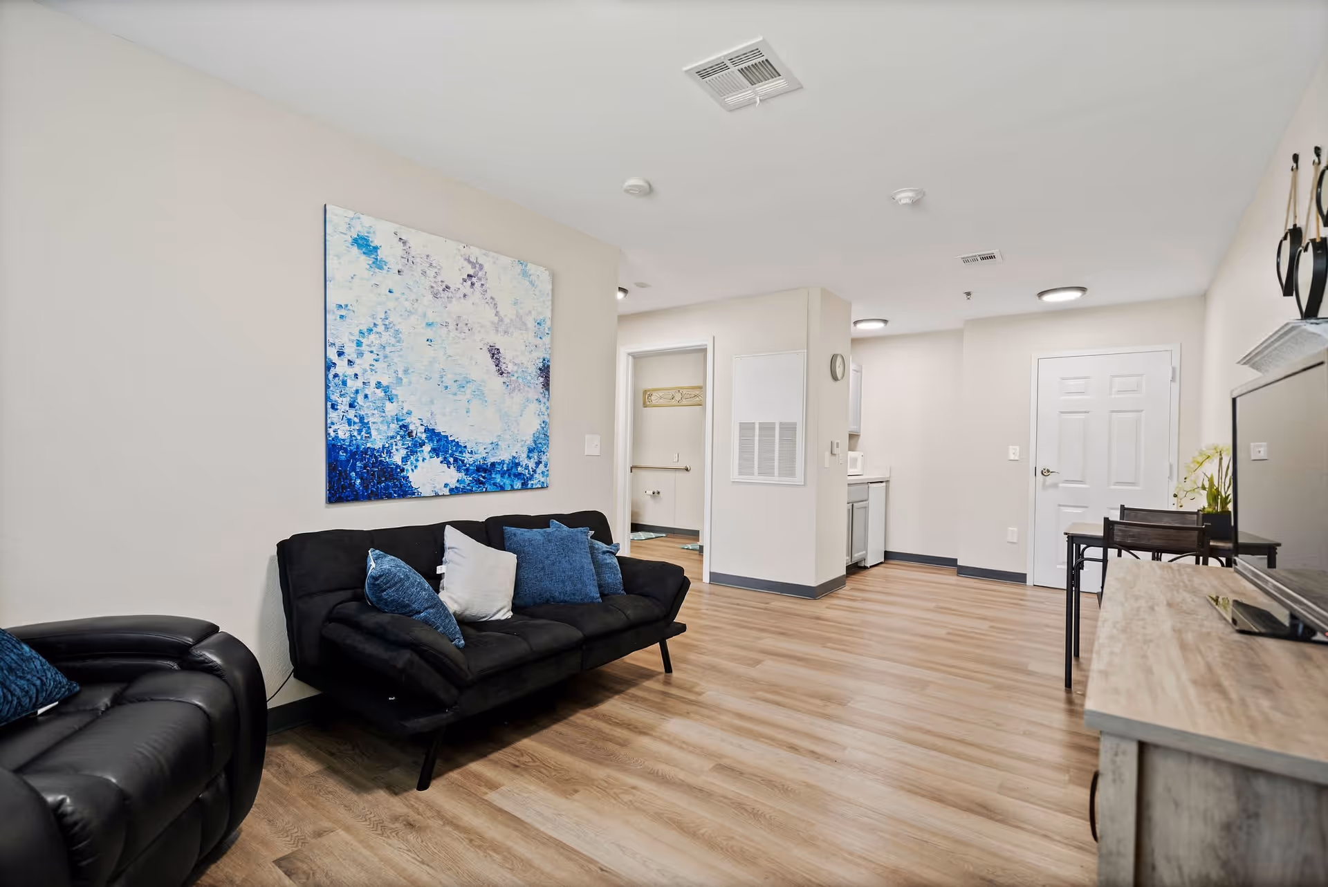 A modern senior living facility interior featuring a black sofa with blue and white cushions, a black recliner chair, a large abstract blue and white painting on the wall, wood flooring, and a small dining area with a table and chairs near a white door. The space is well-lit with ceiling lights and has a clean, minimalist design.