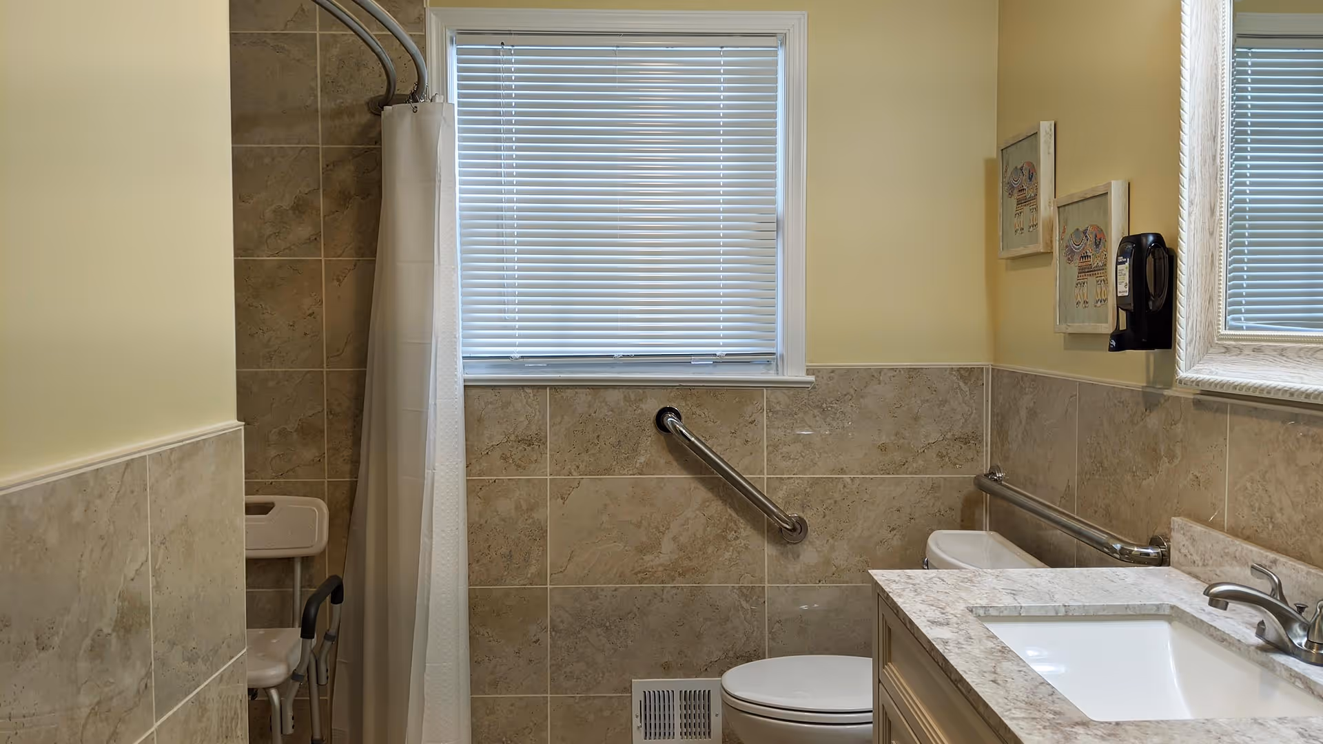 A bathroom with beige tiled walls and a window with closed blinds. There is a white toilet with a grab bar on the wall beside it, a sink with a marble countertop and a faucet, and a shower area with a white curtain and a shower chair. Two framed pictures hang on the wall above a soap dispenser.