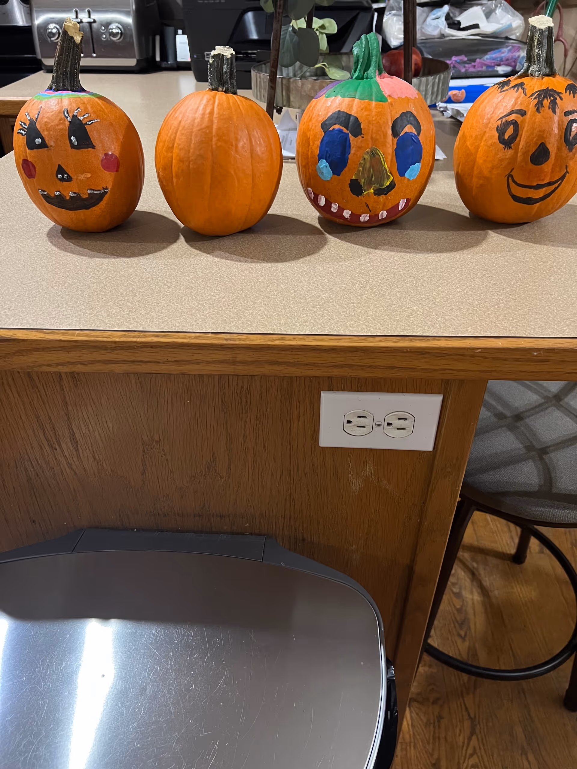 Four small pumpkins lined up on a kitchen counter, three of which are decorated with painted faces. The counter has a wooden front with an electrical outlet, and a metal trash can is visible below. In the background, kitchen appliances and a chair are partially visible.