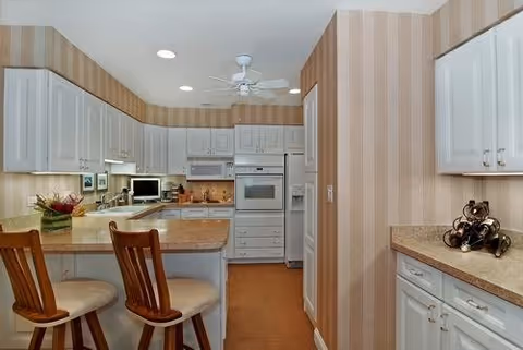 A kitchen with white cabinets, beige countertops, and striped beige wallpaper. There are two wooden chairs with cushioned seats at a counter. The kitchen includes built-in appliances such as an oven and microwave, a ceiling fan, and various kitchen items on the counters.