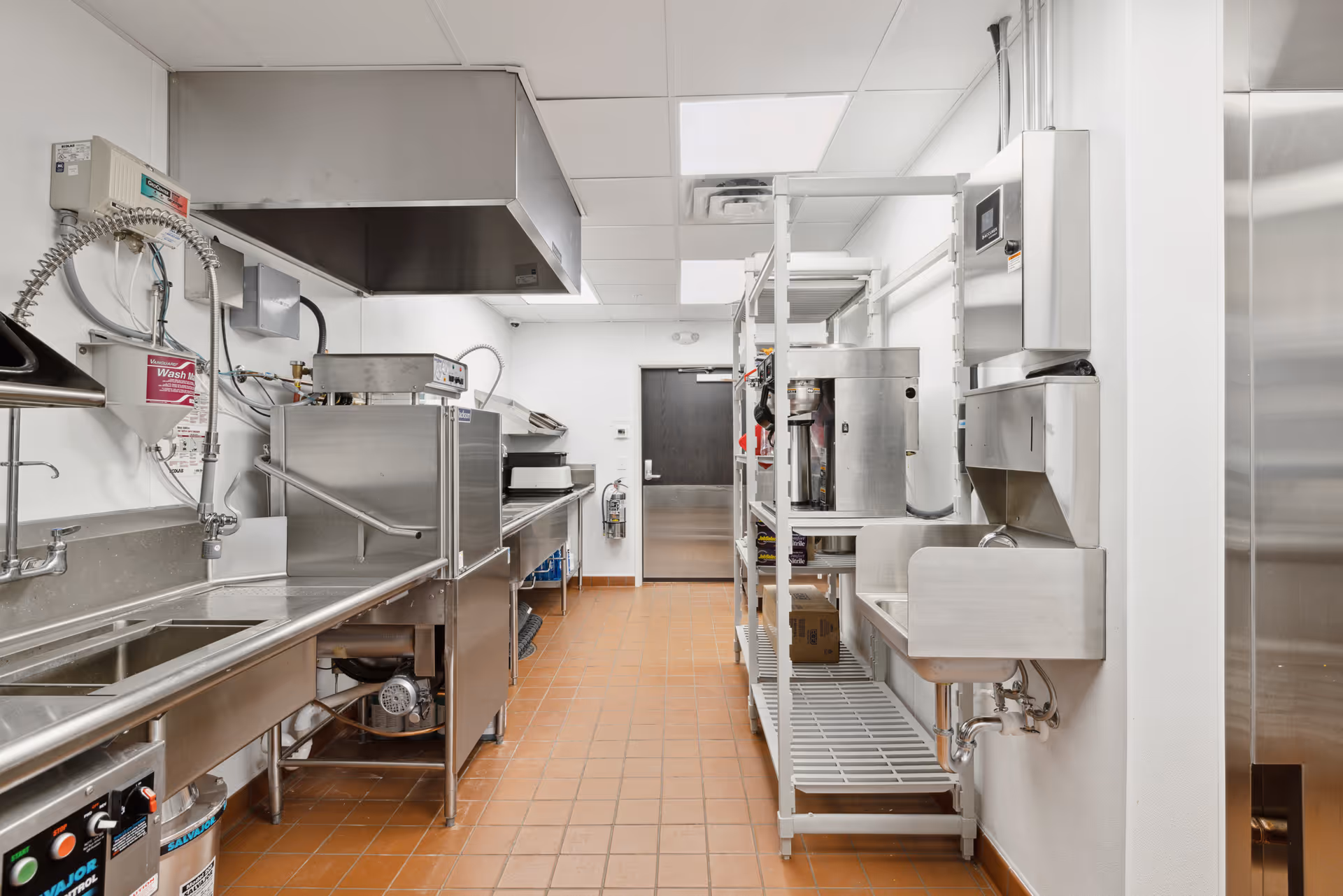 A clean commercial kitchen with stainless steel appliances and fixtures, including a large sink with a flexible spray hose, a dishwasher, metal shelving with kitchen equipment, and a handwashing sink with a soap dispenser mounted on the wall. The floor is tiled with brown tiles and the walls and ceiling are white with fluorescent lighting.