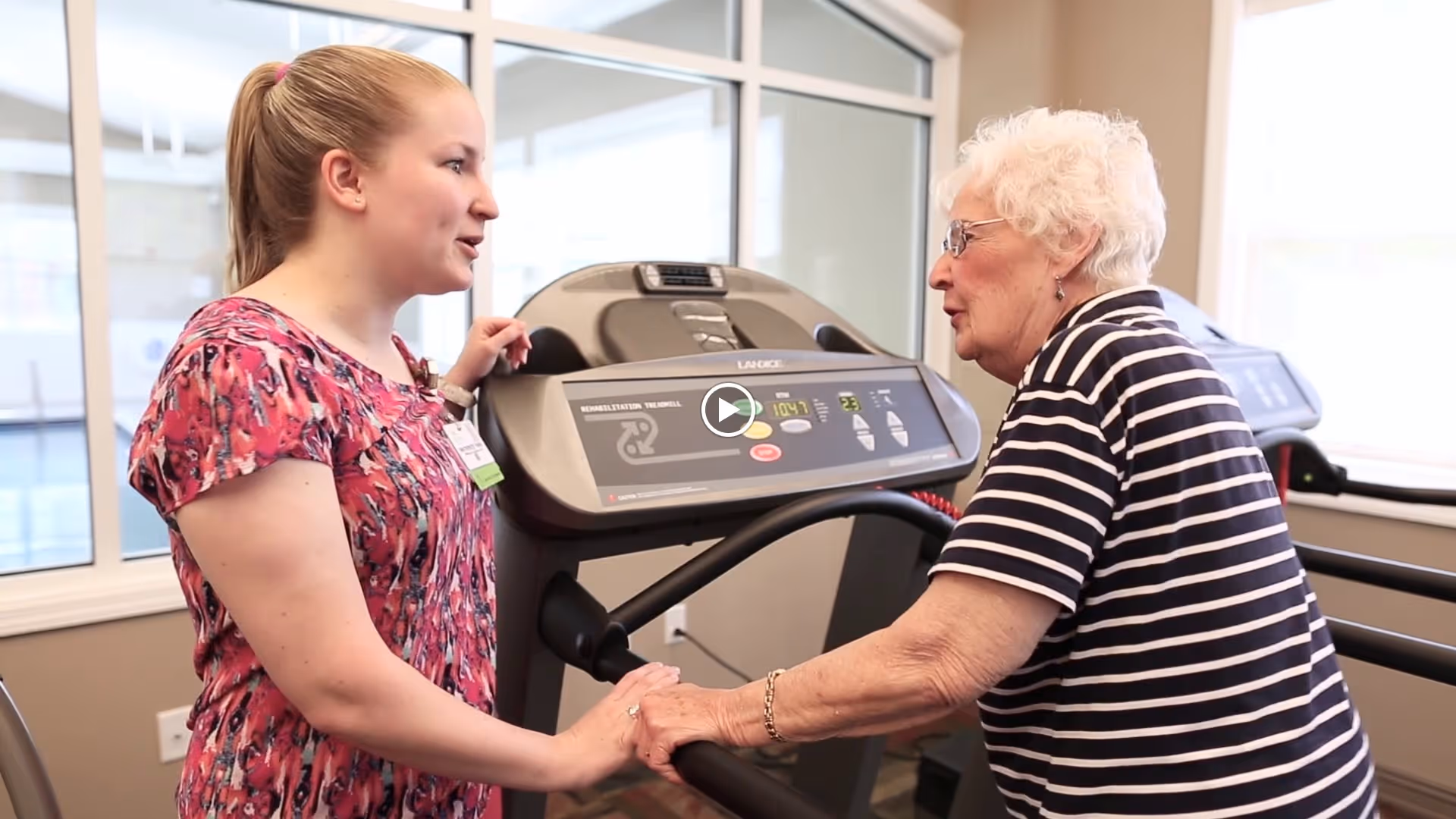 A young woman in a pink patterned shirt is assisting an elderly woman with white hair and glasses who is using a treadmill in a fitness room with large windows.