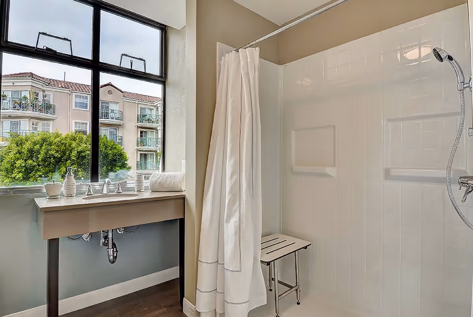 A bright bathroom with a large window showing an outside view of a building and trees. The bathroom features a sink with a countertop holding soap dispensers and a rolled towel. Next to the sink is a shower area with a white curtain, a wall-mounted showerhead, and a small bench inside the shower.