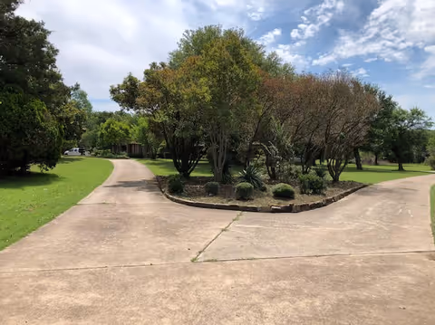 A paved driveway splits into two paths around a landscaped island with trees and shrubs, surrounded by green grass under a partly cloudy sky.