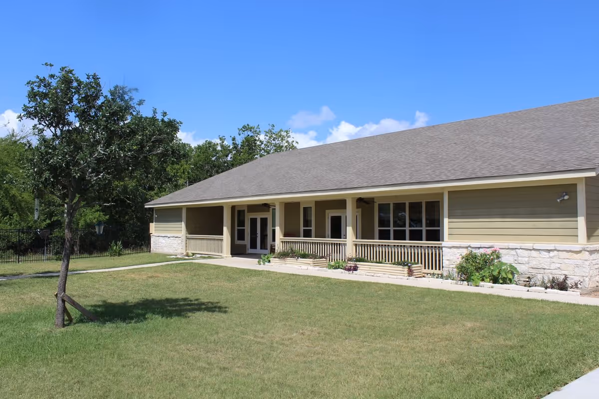 Single-story building with a covered porch, beige siding, and stone accents, surrounded by a grassy lawn and a tree under a clear blue sky.