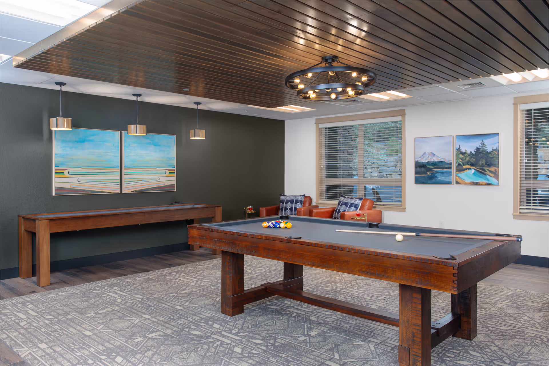 A bright indoor game room with a wooden pool table, seating area, artwork on the walls, and modern overhead lighting.