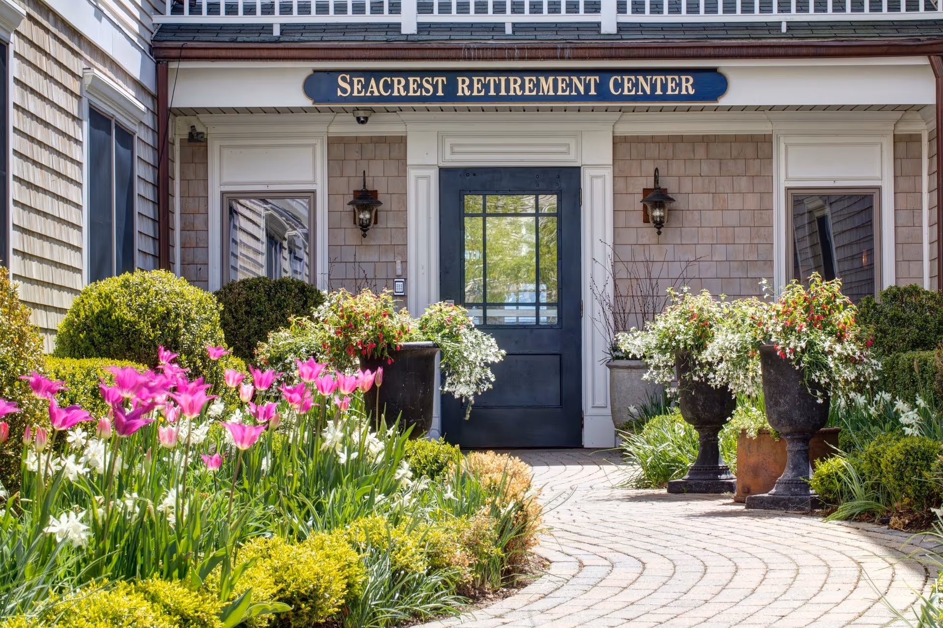Entrance to Seacrest Retirement Center with a black door and windows on either side, surrounded by well-maintained landscaping including pink and white flowers, green shrubs, and large decorative planters. The building exterior features light-colored shingles and two wall-mounted lanterns beside the door.