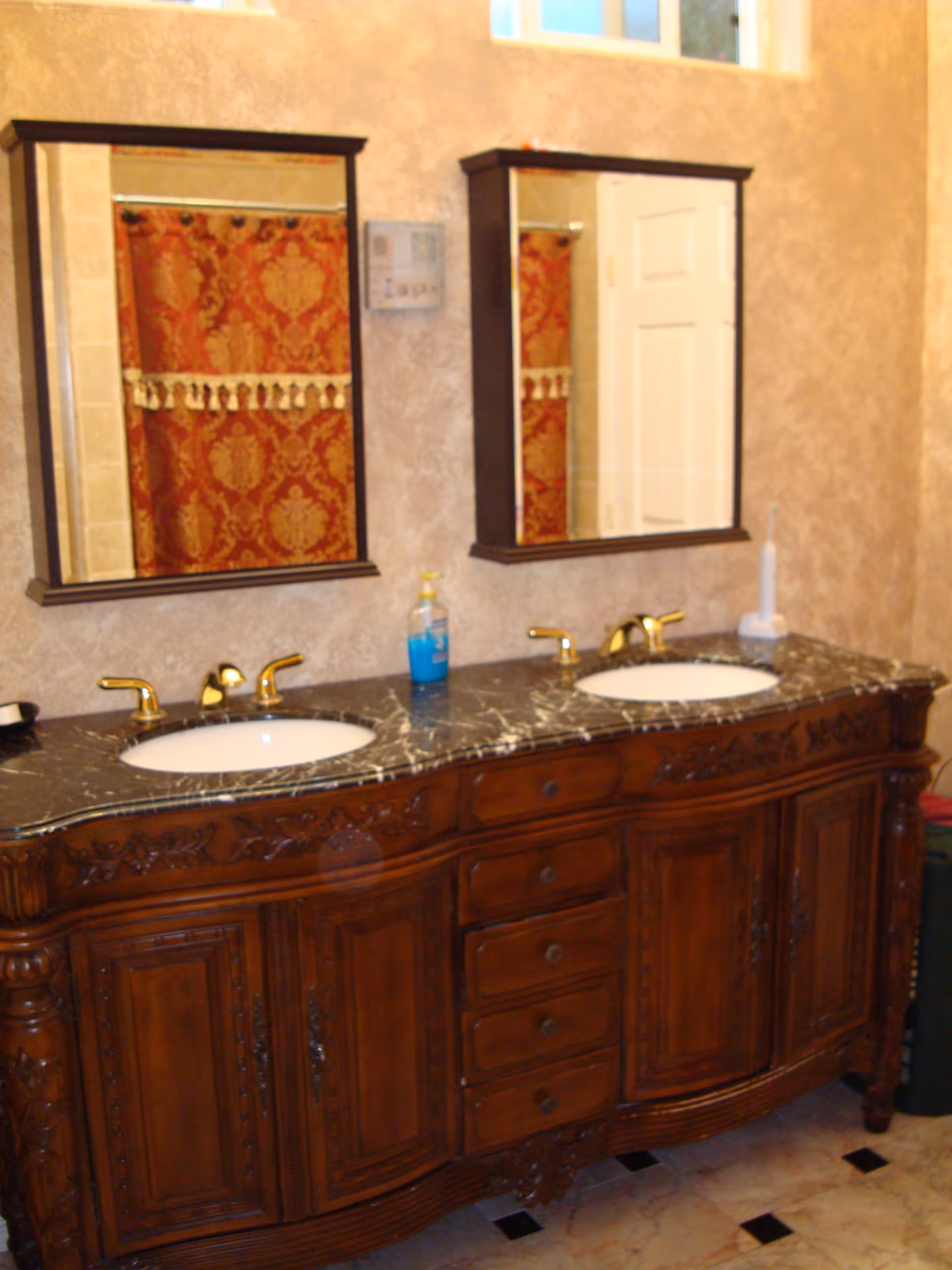A bathroom vanity with a dark marble countertop featuring two white sinks and gold faucets. Above the sinks are two mirrors with dark wooden frames. A red and gold patterned shower curtain is visible in the reflection of the mirrors. The vanity has ornate wooden cabinetry with multiple drawers and doors. The walls have a textured beige finish and the floor is tiled with a light color and black accents.