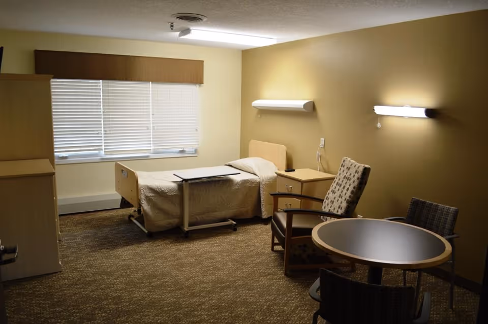 A simple, clean room with a single bed covered with a beige quilt, a small bedside table, a rolling overbed table, a cushioned armchair, a round table with two chairs, and a window with closed blinds and a brown valance. The walls are painted beige and two wall-mounted lights are turned on.