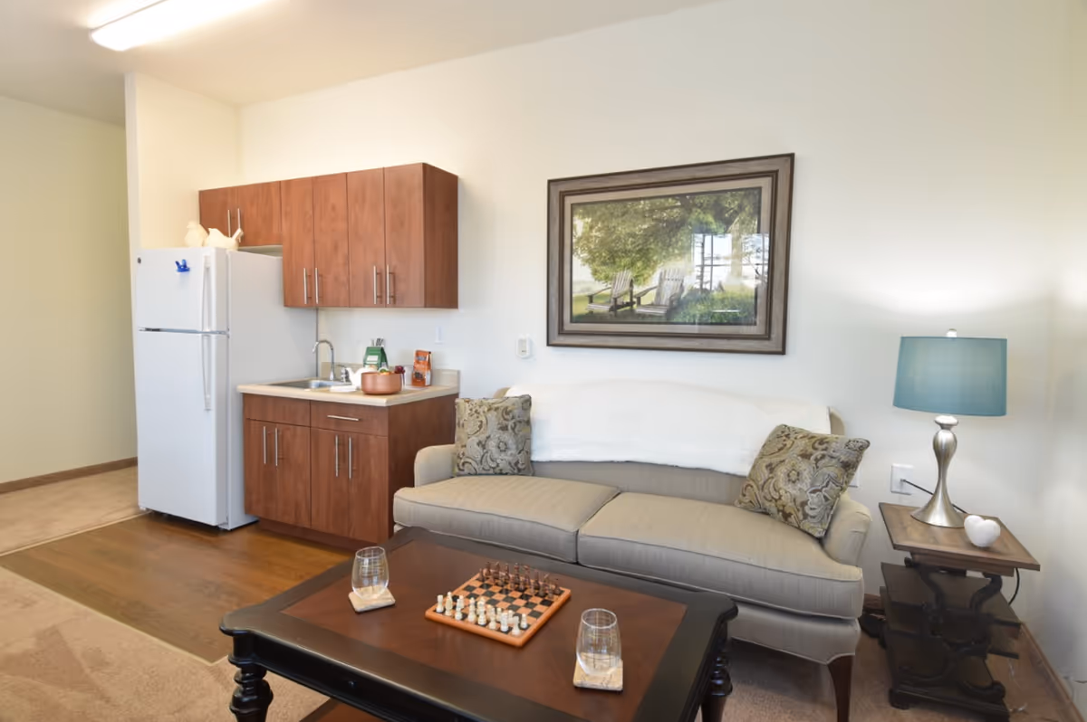 A cozy living area with a beige sofa adorned with two patterned pillows and a white throw blanket. A wooden coffee table in front of the sofa holds a chessboard and two glasses. To the right of the sofa is a side table with a blue lampshade and a small decorative item. On the left side of the room is a compact kitchen area with wooden cabinets, a white refrigerator, and a sink. A framed picture of outdoor chairs under a tree hangs on the wall above the sofa.